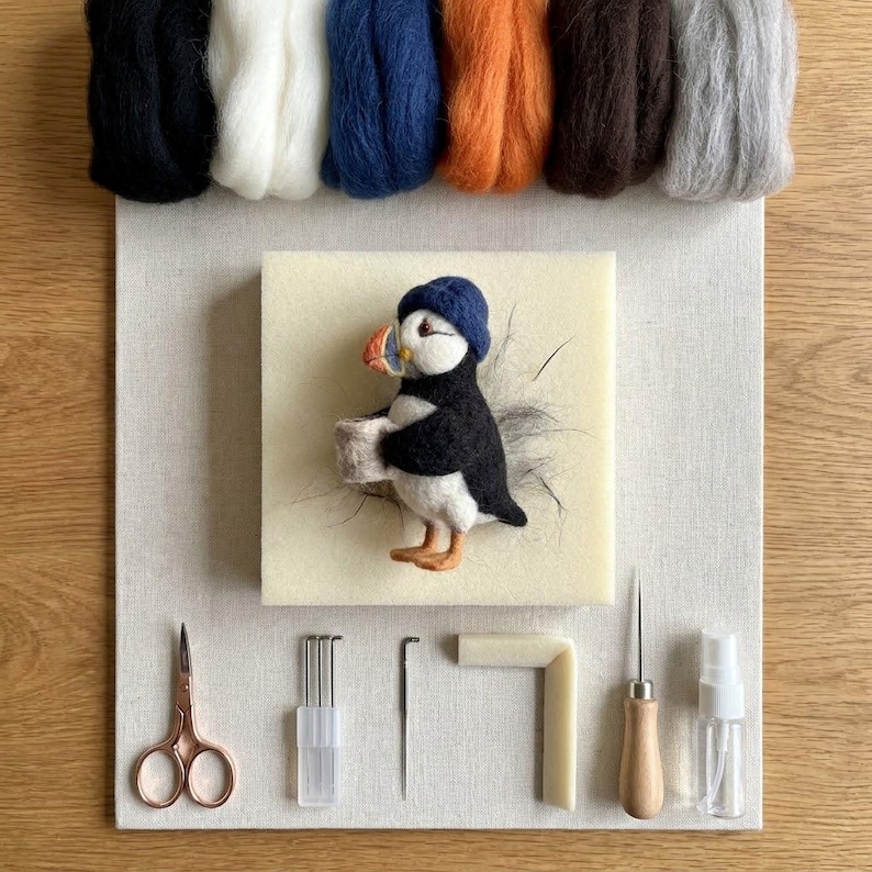 May include: A needle felting kit featuring a puffin figure wearing a blue hat, positioned on a foam block. Included are six bundles of wool roving in black, white, blue, orange, brown, and gray, plus scissors, needles, a tool handle, and a spray bottle.