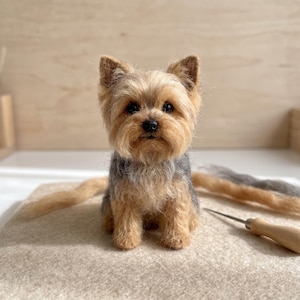 Könnte beinhalten: Eine nadelgefilzte Yorkshire Terrier-Figur mit lohfarbenem und grauem Fell, aufrechten Ohren und schwarzen Perlenaugen. Die kleine Hundeskulptur sitzt in aufrechter Position, begleitet von losen Büscheln aus lohfarbener Wolle und einer Filznadel.
