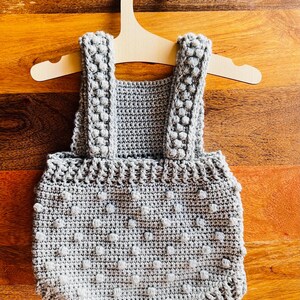 May include: A gray crocheted baby onesie with a textured, dotted pattern. The onesie is hanging on a wooden hanger.