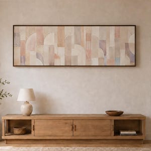 Op de afbeelding: Een lang, rechthoekig wandkunstwerk met een abstract geometrisch patroon in zachte tinten beige, perzik en gedempt blauw, ingelijst in donker hout. Het kunstwerk hangt horizontaal boven een laag mediameubel van lichtgekleurd hout.