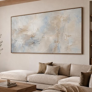 Puede incluir: Una gran pintura abstracta rectangular que presenta una mezcla texturizada de tonos azul suave, crema y beige. La obra de arte está encerrada en un marco de madera delgado y muestra pinceladas en capas que crean profundidad y movimiento.