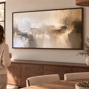 Puede incluir: Una gran pieza de arte mural rectangular que presenta una composición abstracta en tonos beige, crema, marrón y gris carbón. La obra está encerrada en un marco fino de color marrón oscuro y muestra formas y texturas suaves y difusas.