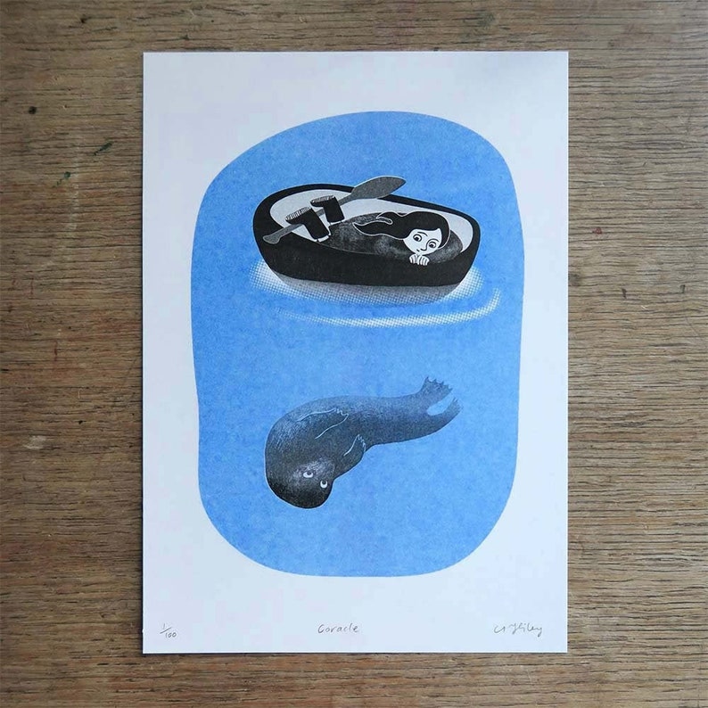 within a round blue shape, a child curled in a coracle rests her head on her hands looking over the edge. underneath a seal floats upside down, looking up at the child. A5 size riso print in blue and black lying on wooden surface.