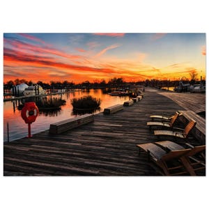 Op de afbeelding: Een houten vlonder bij zonsondergang met diverse bruine ligstoelen, een felrode reddingsboei op een paal en een rustige haven met aangemeerde boten onder een levendige oranje en paarse lucht die reflecteert op het wateroppervlak.
