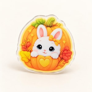 Peut inclure: Une broche en acrylique transparent représentant un lapin blanc de dessin animé avec de grands yeux sombres et un nœud jaune, sortant d'une citrouille orange vif. La citrouille est ornée d'un cœur jaune, de feuilles vertes et de feuillage automnal.