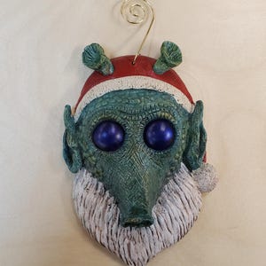 Peut inclure: Une décoration en forme d'extraterrestre vert texturé portant un bonnet de Père Noël rouge à bordure blanche et une barbe blanche. La figurine a deux grands yeux sphériques violet foncé, des oreilles pointues et deux petites tiges sur la tête.