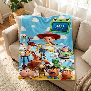 Puede incluir: Una manta de forro polar azul claro con personajes de Toy Story como Woody, Jessie, Buzz Lightyear, Rex, Slinky Dog, Mr. y Mrs. Potato Head, Bullseye, Forky y Hamm. Un marco verde con forma de rana en la parte superior incluye el texto Hi!.