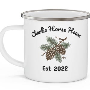 Op de afbeelding: Een witte emaille mok met een zilveren rand en een handvat aan de linkerkant. De voorzijde toont een afbeelding van twee bruine dennenappels op groene dennentakken. Boven de afbeelding staat de tekst Charlie Horse House en onder Est 2022.