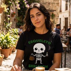 May include: A black short-sleeved t-shirt featuring a graphic of a cartoon skeleton holding a brown paper bag and a lit candle. The text "ESQUELETO" appears in green above the skeleton, and the word "SKELETON" is printed in green below it.