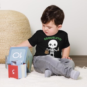 May include: A black short-sleeved t-shirt featuring a graphic of a cartoon skeleton holding a brown paper bag and a lit candle. Above the skeleton, the word ESQUELETO is printed in green capital letters. The shirt is paired with grey trousers.