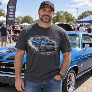 May include: A charcoal gray short-sleeve t-shirt featuring a graphic print of a blue muscle car with black racing stripes surrounded by white tire smoke. The shirt is worn with blue jeans and a dark baseball cap with an American flag patch.
