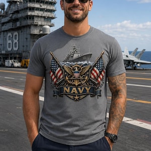 以下が含まれることがあります： 軍艦、交差した2つのアメリカ国旗、そして中央に鷲、錨、NAVYの文字が書かれたバナーが描かれたエンブレムがプリントされたグレーのTシャツ。デザインには、詳細なゴールドとブルーのアクセントが含まれています。