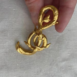 Op de afbeelding: Een goudkleurige broche met een abstract, verstrengeld ontwerp met getextureerde oppervlakken en kleine, puntige, bladachtige uitsteeksels. Het metalen stuk heeft een matte, geborstelde afwerking en een continue, organische lusstructuur.
