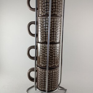 May include: A set of four brown ceramic mugs with a textured, grid-like pattern stacked vertically in a chrome metal wire stand. Each mug features a rounded handle on the side, and the stand has a square base with four vertical support rods.