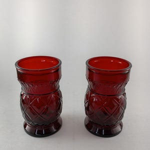 May include: Two matching ruby red glass tumblers featuring a textured diamond pattern on the lower body and a smooth, slightly flared rim. The glassware has a cylindrical shape with a thick base and a distinct embossed design around the upper section.