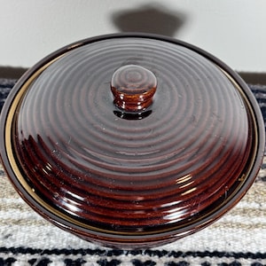 May include: A round, dark brown ceramic lidded bowl featuring a glossy finish and a series of concentric circular ridges across the top of the lid. The lid includes a small, centered knob handle, and the bowl has a smooth, reflective surface.