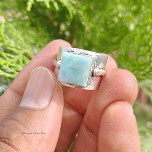May include: A silver ring featuring a square, light blue, marbled stone set in the center. The band has a wide, textured, metallic finish with small decorative silver spheres on either side of the stone setting. The text MoonstoneSilverStore is visible.