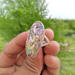May include: An oval-shaped ring featuring a large, iridescent abalone shell inlay with swirling patterns of pink, green, and blue hues, set within a polished silver metal band that splits into two distinct arms at the sides of the setting.