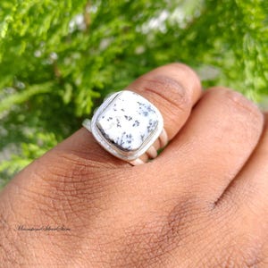 May include: A silver ring featuring a square-shaped dendritic opal gemstone with a white base and black fern-like inclusions. The stone is set in a smooth silver bezel on a simple silver band. The text MoonstoneSilverStore is visible at the bottom.