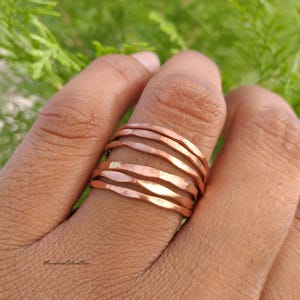 Könnte beinhalten: Ein Set aus fünf dünnen, gestapelten Ringen aus poliertem Kupfer. Jeder Ring weist eine leicht gehämmerte, unebene Textur entlang der Ringschiene auf, was ein wellenförmiges Aussehen erzeugt. Die Ringe werden zusammen an einem Finger getragen.
