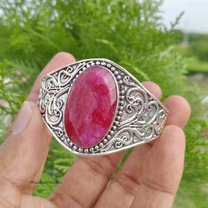 May include: A silver cuff bracelet featuring a large, oval-shaped, faceted deep pink gemstone at the center. The metal band is decorated with intricate, raised scrollwork patterns and a beaded border surrounding the central stone setting.