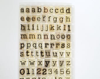 May include: A clear sheet of individual photopolymer stamps featuring lowercase letters, numbers, and punctuation marks in a typewriter-style font. The set includes multiple instances of common letters, digits zero through nine, and symbols like ! ? , . ' & #.