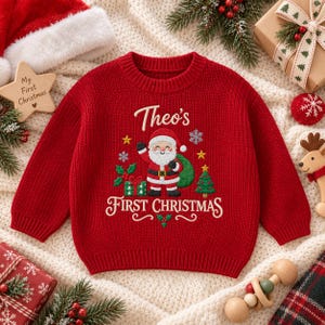 May include: A red long-sleeve knit sweater featuring an embroidered Santa Claus, a green gift, a Christmas tree, and snowflakes. The text "Theo's FIRST CHRISTMAS" is embroidered in white across the front. The sweater has a ribbed crew neckline.