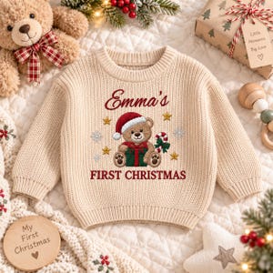 May include: A beige long-sleeve knit sweater featuring an embroidered brown teddy bear wearing a red Santa hat, holding a green gift box with a red bow, and a candy cane. The text reads Emma's FIRST CHRISTMAS with gold stars and snowflakes.