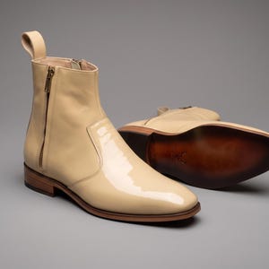Könnte beinhalten: Ein Paar knöchelhohe Stiefel aus cremefarbenem Lackleder mit seitlichem Reißverschluss, einer Zugschlaufe an der Ferse und einem geschichteten Holzabsatz. Die Sohle des zweiten Stiefels ist sichtbar und zeigt ein dunkelbraunes Finish mit Logo.
