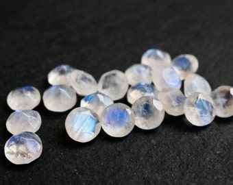 May include: A group of 15 iridescent moonstone cabochons, each with a unique blue sheen, are scattered on a black surface. The stones are smooth and polished, with a slightly rounded shape.