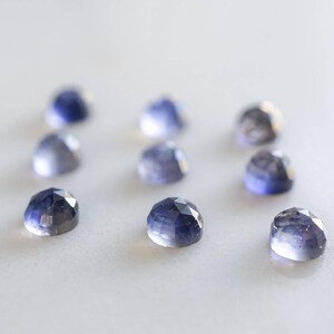 May include: A collection of eight faceted, blue-grey gemstones on a white surface. The gemstones are all approximately the same size and shape.