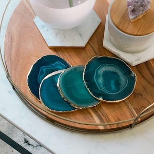 Könnte beinhalten: Vier tealfarbene Achat-Untersetzer mit goldener Einfassung auf einem Holztablett mit einer weißen Marmorplatte. Eine klare Glasvase mit rosa Sockel und einer grünen Pflanze steht auf dem Tablett. Ein weißer Keramiktopf mit einem Holzdeckel und einem violetten Kristall darauf steht ebenfalls auf dem Tablett.
