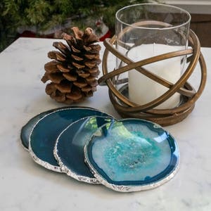 May include: Four teal blue agate coasters with silver trim. The coasters are arranged in a stack on a white marble surface.