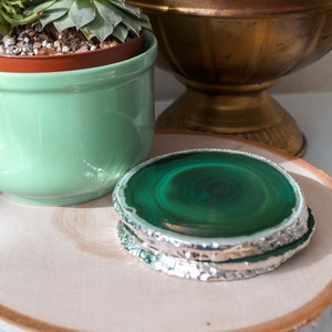May include: A green agate coaster with a silver trim sits on a wooden surface. The coaster is round and has a natural, polished finish.