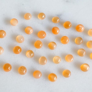 Puede incluir: Una colección de 25 piedras preciosas redondas de cabujón de color naranja. Las piedras son lisas y tienen un ligero brillo.