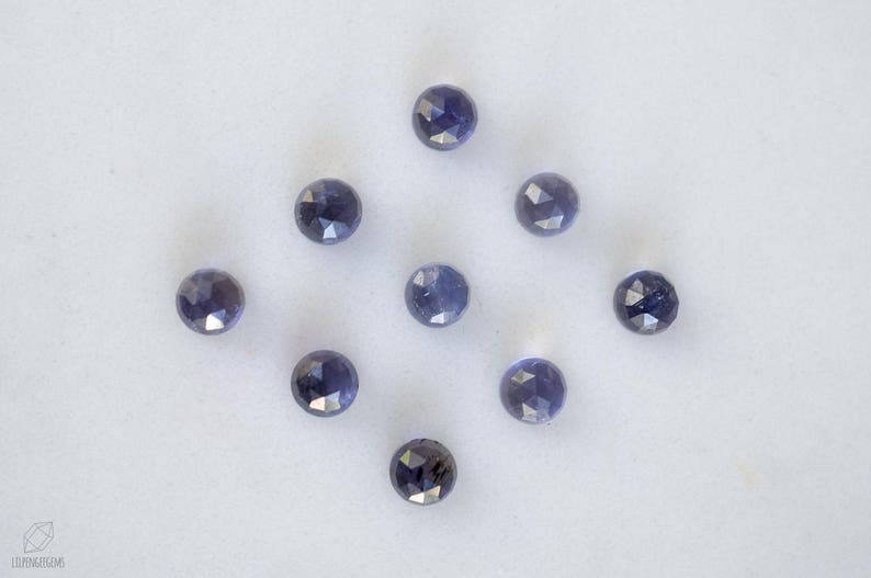 May include: Ten faceted, round, dark blue gemstones on a white surface.