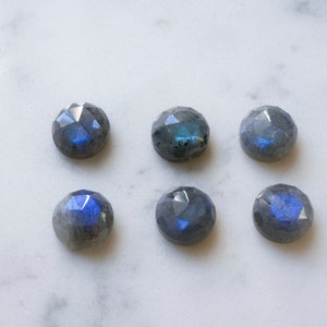 Puede incluir: Seis piedras preciosas redondas y facetadas de labradorita de color gris oscuro con destellos azules iridiscentes. Las piedras están dispuestas en dos filas de tres sobre una superficie de mármol blanco.