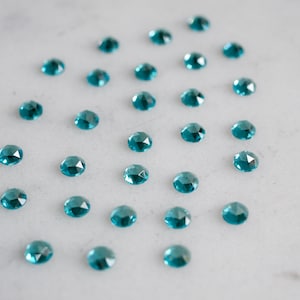 May include: A collection of teal blue faceted gemstones arranged in a pattern on a white marble surface.