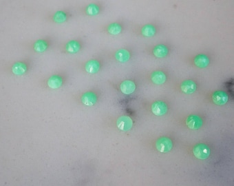 Pode incluir: Uma coleção de 20 cabochões de pedras preciosas verdes, cada um com uma forma facetada única. Os cabochões estão dispostos sobre uma superfície branca.