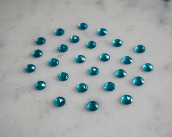 May include: A collection of teal blue faceted gemstones arranged in a circle on a white marble surface.