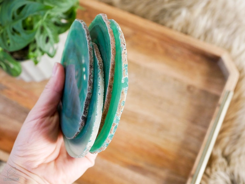 May include: A set of four green agate coasters with a natural, raw edge. The coasters are stacked on top of each other and are being held in a hand.