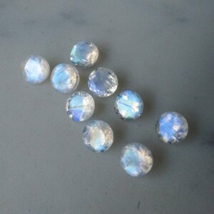 May include: A collection of nine round, faceted moonstone gemstones. The stones display a milky white base color with iridescent blue and white flashes. They are scattered on a gray surface.