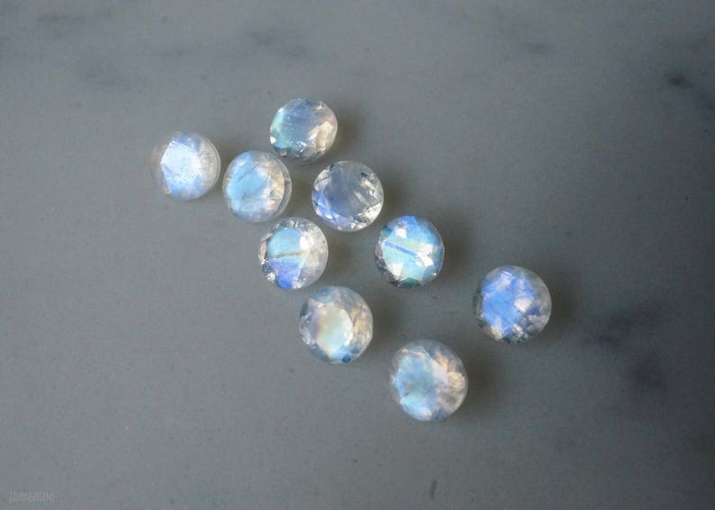 May include: A collection of nine round, faceted moonstone gemstones. The stones display a milky white base color with iridescent blue and white flashes. They are scattered on a gray surface.
