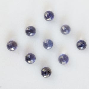 May include: Nine faceted, round, purple gemstones on a white surface.