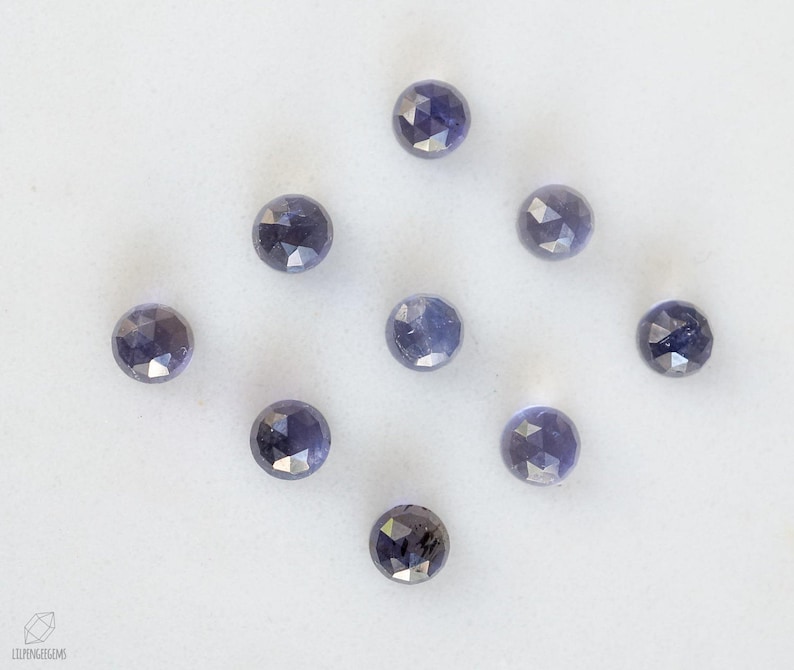 May include: Nine faceted, round, purple gemstones on a white surface.