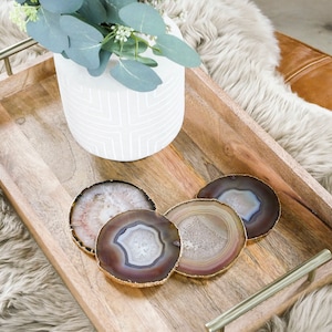 Puede incluir: Una bandeja de madera con asas doradas, con cuatro posavasos de ágata. Los posavasos son redondos y tienen un patrón natural marrón y blanco. La bandeja está sobre una alfombra blanca y esponjosa.