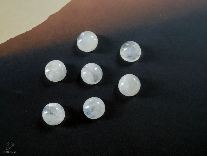 Puede incluir: Siete cabujones redondos de piedra lunar blanca, cada uno con un patr&oacute;n de inclusiones ligeramente diferente, est&aacute;n dispuestos en c&iacute;rculo sobre una superficie marr&oacute;n oscura.