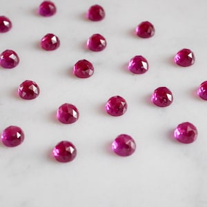 Peut inclure: Une collection de 20 pierres précieuses de rubis facettées, chacune avec une teinte rose foncé, disposées en grille sur une surface blanche.
