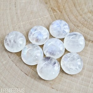 Puede incluir: Ocho cabujones de piedra lunar blanca lisos con brillo iridiscente. Las piedras est&aacute;n dispuestas en c&iacute;rculo sobre una superficie de madera marr&oacute;n claro.