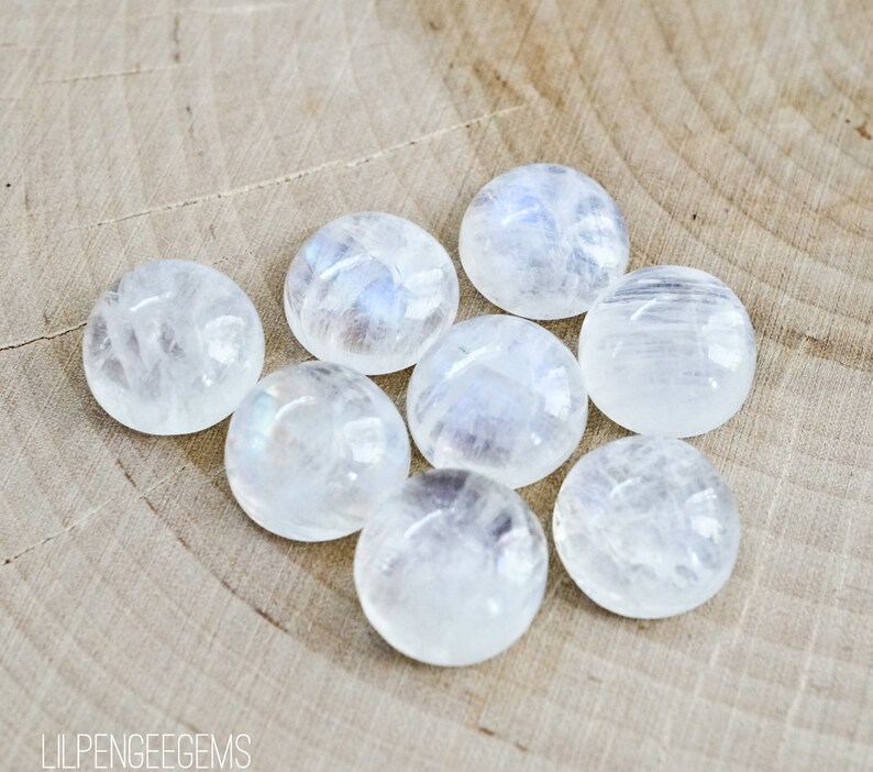 Puede incluir: Ocho cabujones de piedra lunar blanca lisos con brillo iridiscente. Las piedras est&aacute;n dispuestas en c&iacute;rculo sobre una superficie de madera marr&oacute;n claro.